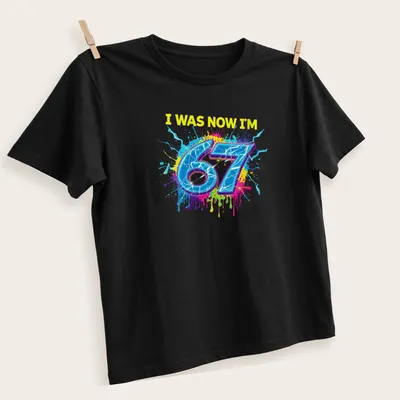 Colorful splatter design t-shirt with the text I WAS NOW I'M 67 — angled view
