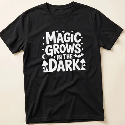 A dark themed t-shirt with the text 'Magic Grows In The Dark' surrounded by plant illustrations in black, green, and purple.